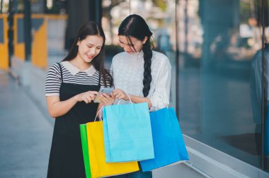 İki kadın bir mağazanın önünde cep telefonlarına bakıyor. Alışveriş çantalarını tutuyorlar ve telefonlarından bir şeyi kontrol ediyor gibi görünüyorlar.