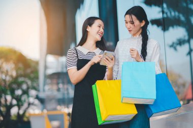 İki kadın bir mağazanın önünde duruyor, biri cep telefonu tutuyor. İkisi de gülümsüyor ve alışveriş deneyimlerinden zevk alıyor gibi görünüyorlar.