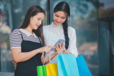 İki kadın alışveriş torbaları tutarken cep telefonuna bakıyor. Telefondaki bir şeyi kontrol ediyorlar gibi görünüyor, muhtemelen bir alışveriş listesi ya da kupon. Kolaylık ve modernlik kavramı