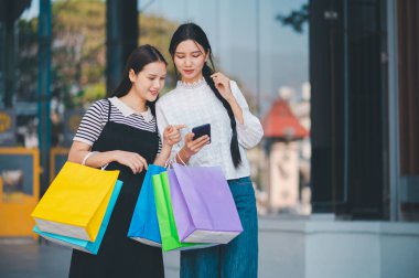 İki kadın ellerinde alışveriş torbalarıyla cep telefonlarına bakıyor. Sahne rahat ve rahattır, çünkü kadınlar alışveriş gezilerinin tadını çıkarıyorlar ve telefonlarını kontrol etmek için mola veriyorlar.