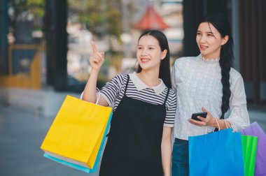 İki kadın alışveriş yapıyor ve biri cep telefonunu işaret ediyor. Diğer kadın bir çanta tutuyor ve ikisi de gülümsüyor.