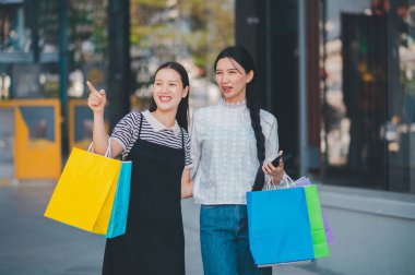 İki kadın sokakta yürüyor, her biri bir alışveriş çantası tutuyor. Kadınlardan biri uzaktaki bir şeyi işaret ediyor.
