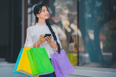 Bir kadın alışveriş torbalarını tutuyor ve telefonuna bakıyor. Gülümsüyor ve alışveriş deneyiminin tadını çıkarıyor.