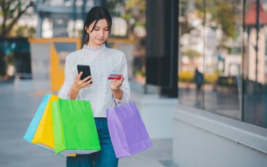Bir kadın elinde bir çanta ve cep telefonuyla sokakta yürüyor. Alışveriş çantalarını tutarken telefonuna bakıyor.