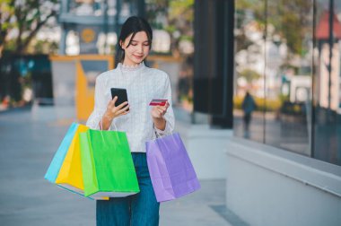Bir kadın elinde cep telefonu ve alışveriş torbalarıyla sokakta yürüyor.