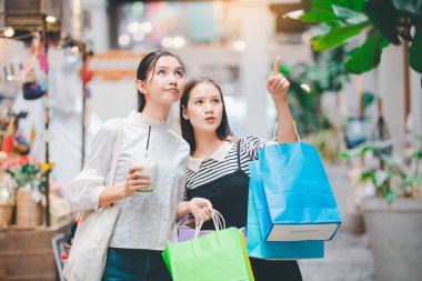 İki kadın alışveriş merkezinde alışveriş yapıyor, biri bir mağazayı işaret ediyor. Birden fazla çanta taşıyorlar, biri mavi olmak üzere.