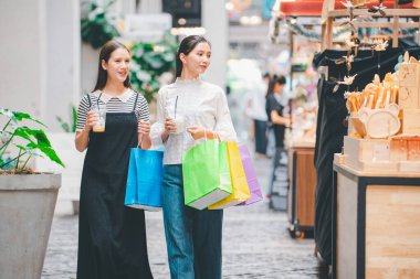 İki kadın ellerinde alışveriş çantalarıyla sokakta yürüyorlar. Kadınlardan biri yeşil bir çanta tutuyor.