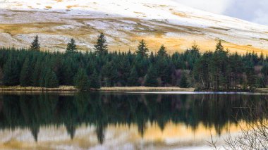 Kış manzarası ve yansımaları Brecon Beacons Ulusal Parkı 'ndaki Galler rezervuarının suyunda kar ve buzla kaplı buzlu dağ tarafını ve yukarıda bulutlu gökyüzünü gösteriyor.. 