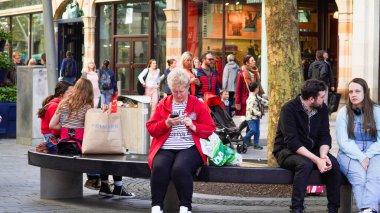 Cardiff, Wales, 29 Ekim 2023: Sokak fotoğrafçılığı, Cardiff şehir merkezinde yoğun bir öğleden sonra. İnsanlar dinlenmek için banklarda otururlar ve son sınıf bir kadın akıllı telefonlarında mesaj arar.