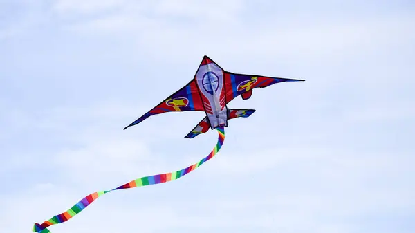 stock image Rocket shaped kite, soaring through the blue sky with white clouds in places. The movement of the long striped multicoloured tail creates pattern and shape as twists and turns. With copy space.