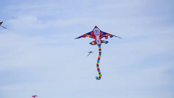 stock image Rocket shaped kite, soaring through the blue sky with white clouds in places. The movement of the long striped multicoloured tail creates pattern and shape as twists and turns. With copy space.