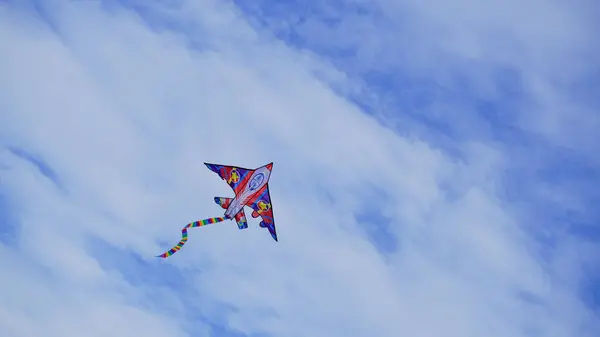 stock image Rocket shaped kite, soaring through the blue sky with white clouds in places. The movement of the long striped multicoloured tail creates pattern and shape as twists and turns. With copy space.