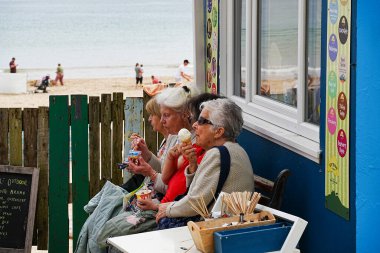 Tenby, Pembrokeshire, Galler - 2 Temmuz 2024: Güney sahilinin yanındaki bankta oturan dört son sınıf öğrencisi birlikte dondurma yiyorlar.