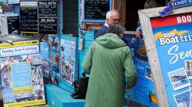 Tenby, Pembrokeshire, Galler - 02 Temmuz 2024: Limanın girişinde balıkçılık ve gezi gezileri satan küçük kulübeler bulunmaktadır. Parlak mavi işaretler canlı, göz alıcı ve bilgilendiricidir..