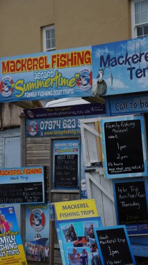 Tenby, Pembrokeshire, Galler - 02 Temmuz 2024: Limanın girişinde balıkçılık ve gezi gezileri satan küçük kulübeler bulunmaktadır. Parlak mavi işaretler canlı, göz alıcı ve bilgilendiricidir..