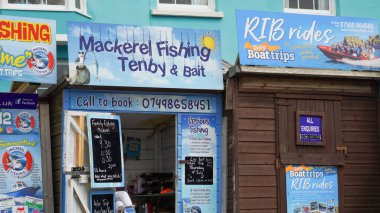 Tenby, Pembrokeshire, Galler - 02 Temmuz 2024: Limanın girişinde balıkçılık ve gezi gezileri satan küçük kulübeler bulunmaktadır. Parlak mavi işaretler canlı, göz alıcı ve bilgilendiricidir..