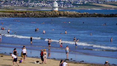 Porthcawl, Bridgend, Wales - August 16 2024: Crowds of people on Coney Beach in Porthcawl enjoying the hot August sun. Children play on the sand and bathe in the sea with the old lighthouse behind. clipart