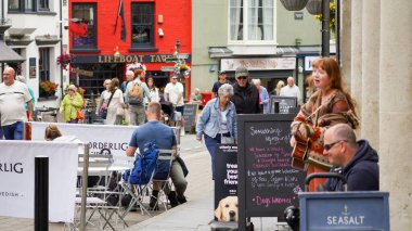 Tenby, Pembrokeshire, Galler - 01 Temmuz 2024: Kızıl saçlı genç bir bayan müzisyen, Tenby şehir merkezinin ana caddesinde yerli halkı ve turistleri eğlendiriyor.