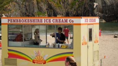 Tenby, Pembrokeshire, Wales - July 03 2024: An ice cream kiosk on the sand at Castle Beach in Tenby with St Catherine's Island towering behind just off the shoreline. clipart