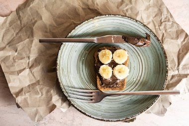 top view of homemade chocolate toast with chocolate cream and banana in a plate, knife and fork, on the table, crumpled paper