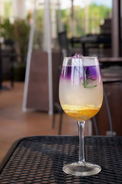 colorful gourmet craft cocktail made with purple gin garnished with a flower and orange slice with soft focus outdoor patio background