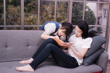 Gülümseyen Asyalı anne ve küçük kız çocuğu oturma odasında kanepede birlikte kitap okuyor. Ev okulu ve eğitim konsepti