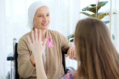 Gülümseyen Kafkasyalı genç kız kemoterapiden sonra tekerlekli sandalyede oturan beyaz başörtülü Yaşlı Anne ile el ele tutuşuyor. Kanser ya da lösemi hastası. Ellere odaklan.
