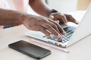 Afrikalı bir işadamının ellerine kapanıp modern ofisteki ahşap masada çevrimiçi toplantı için dizüstü bilgisayarda klavye çalıyor ve daktilo kullanıyor. Akıllı telefon kavramına sahip iş ve teknoloji