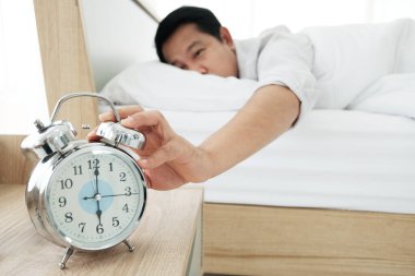 Asyalı bir adam geç kalkar ve sabah yatağın alarmını kapatır ve işe gitmek için tembellik eder. O hasta ve tatilde yorgun hissediyor. El ve saate odaklan.