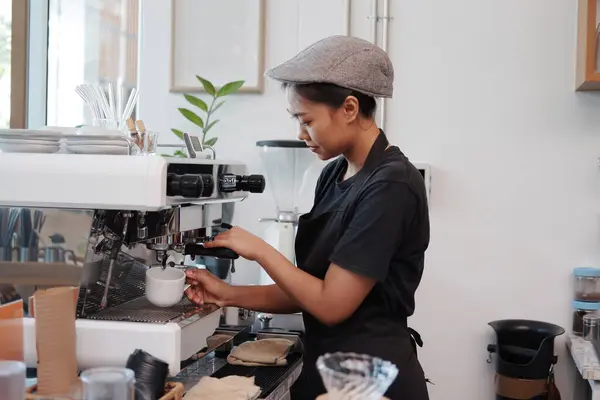 Smiling Asian Young barista woman is wearing apron and brewing coffee with machine for customers order in cafe and coffee shop. Start up small cafe business and technology Concept