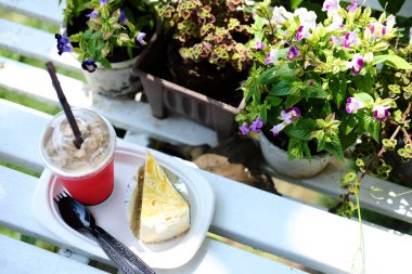 Taze buzlu kahve kırmızı bardakta, peynirli kek ve kremalı ahşap bankta, kafenin çiçek bahçesinde ve kahve dükkanında.