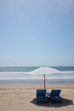 Açık havada kumsalda beyaz şemsiyenin altında iki ahşap mavi koltuk ve güneş ışığında mavi deniz. Endonezya 'da Bali Okyanusu