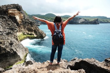 Asyalı kadın sırtı dönük duruyor ve kollarını özgürlük için kaldırıyor sakin denizin kayalık uçurumunda. Varış yeri sırt çantası yazın Endonezya 'daki Bali adasında okyanusla birlikte seyahat eder.