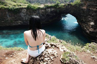 Beyaz şortlu Asyalı kadın, Endonezya 'nın Bali adasındaki kırık plajda, mutlu ve varış noktası okyanus olan kayalık uçurumda sırtüstü oturuyor.