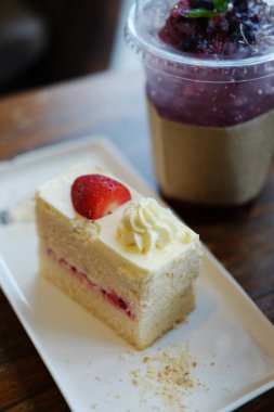 Bir parça vanilyalı kek taze pişmiş çilekli pasta beyaz tabakta ev yapımı kremalı ve yaban mersinli buzlu soda kafedeki ahşap masada plastik cam içinde.