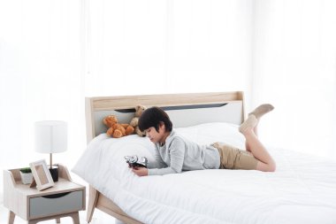 Asyalı tembel bir çocuk çalar saati tutuyor ve sabah beyaz yatakta gürültüyle uyanıyor. Tatilde yatak odasında.