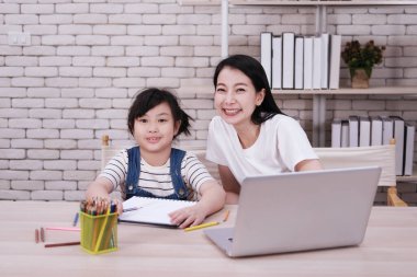 Gülümseyen Asyalı anne ve kız evdeki oturma odasında ahşap masaya renkli kalemle resim yapıyorlar. Evde eğitim kavramı için çocuk eğitimi