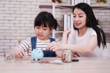 Gülümseyen Asyalı kız ve anne kütüphanedeki ahşap masaya para biriktirmek için kumbaraya bozuk para koyuyorlar. Evde eğitim kavramı için çocuk eğitimi