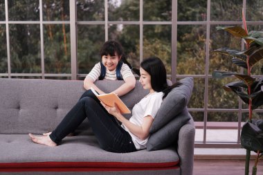 Gülümseyen Asyalı anne, kızına evdeki kanepede kitap yazmayı öğretiyor. Evde eğitim kavramı için çocuk eğitimi