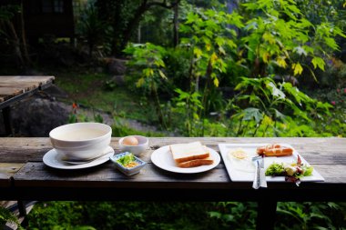 Taze sosisli ve haşlanmış pirinçli sahanda yumurta ve tereyağlı tost. Sabah kahvaltısı için ahşap masada. Tayland 'daki tropik tatil köyünün bahçesinde.
