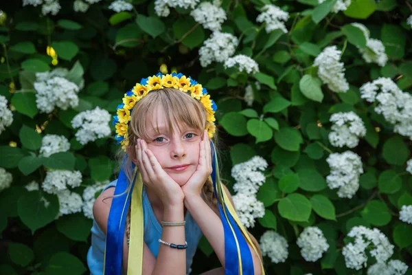 開花茂みの背景にウクライナの旗の色のリボンと青黄色のウクライナの花輪の女の子 — ストック写真