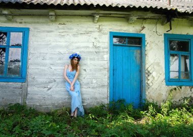 Red-haired girl in a blue wreath near an old wooden house with wooden blue frames.