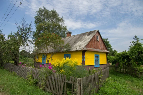 A bright rural wooden house of yellow color behind a fence among flowers against a blue sky.