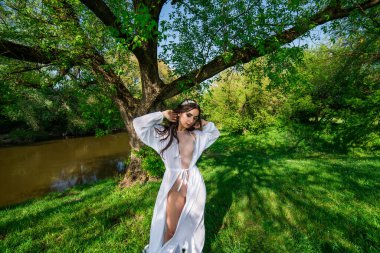 A girl in a bathing suit and a white cape near a broken tree among greenery.