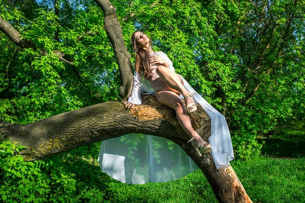 A girl in a bathing suit and a white cape near a broken tree among greenery.
