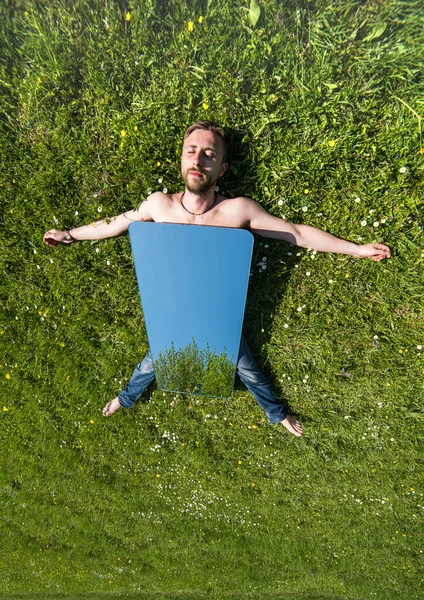 A man with a mirror lies on a green meadow grass, spreading his arms to the sides.