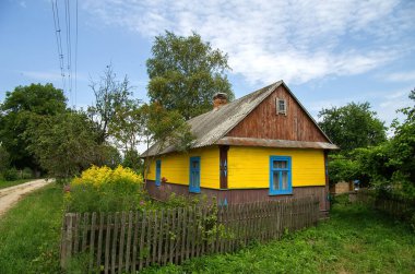 A bright rural wooden house of yellow color behind a fence among flowers against a blue sky.