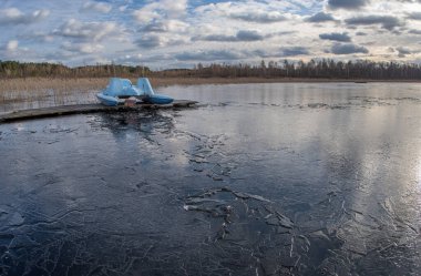 Evening landscape with a water catamaran on a frozen spring lake covered with thin ice with the sky reflected on the surface.