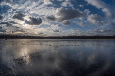 Evening landscape of a spring lake covered with thin cracking transparent ice and a cloudy sky reflected in it. Fish eye.