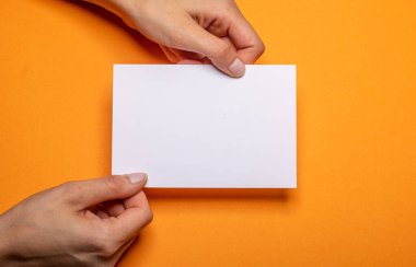 two female hands holding a blank piece of paper with copy space in front of a orange background cardboard